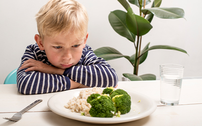 Young boy in striped shirt frowning at kitchen table with arms folded staring at a plate with broccoli and rice.